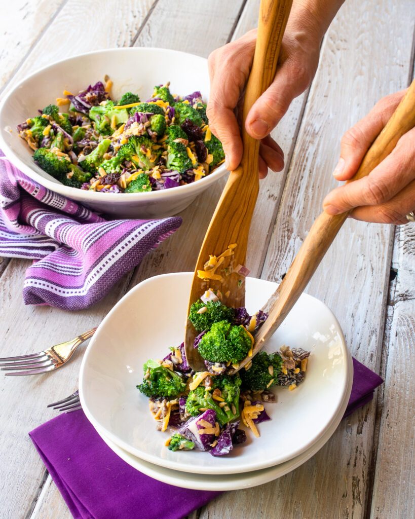 Hands serving broccoli salad into a white bowl with a purple napkin underneath.