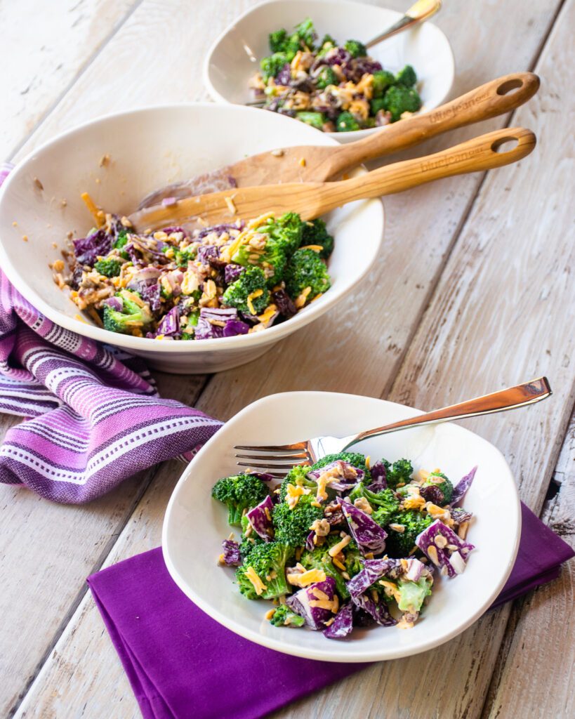 A bowl of broccoli salad on a white wooden table with purple napkin, and the serving salad bowl in the background.