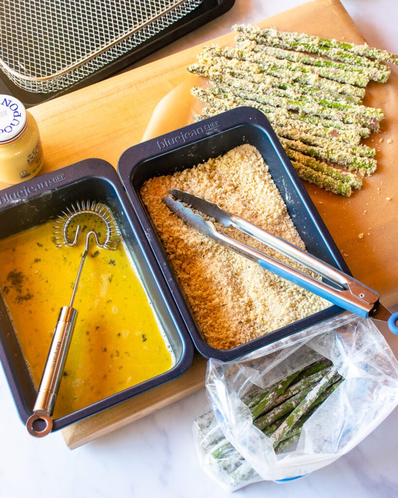 Asparagus being dredged. Asparagus tossed with flour in a plastic bag, a blue tray with beaten egg, a blue tray with breadcrumbs, a pair of tongs and an air fryer basket.