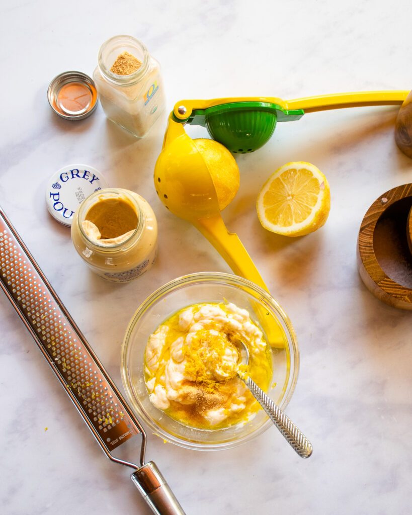 Lemon Aioli ingredients on a marble countertop, ready to be stirred together. Dijon mustard, garlic powder, lemon halves and a bowl with mayonnaise, lemon zest and oil near by.