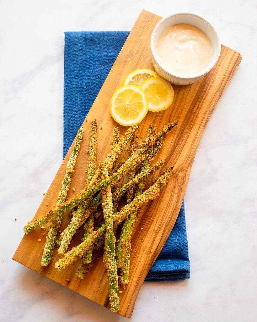 Breaded asparagus on a wooden board with a ramekin of lemon aioli on top of a blue napkin.