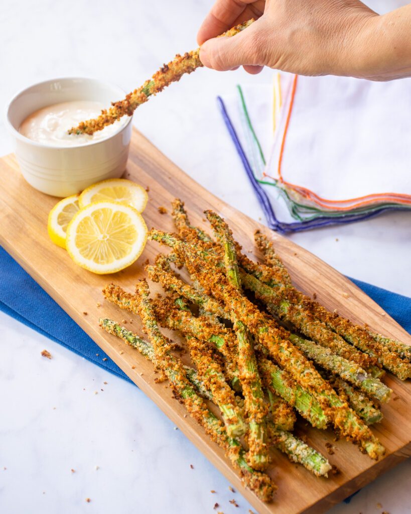 A hand dipping breaded asparagus into a ramekin of lemon aioli with the rest of the breaded asparagus on a wooden board with lemon slices.