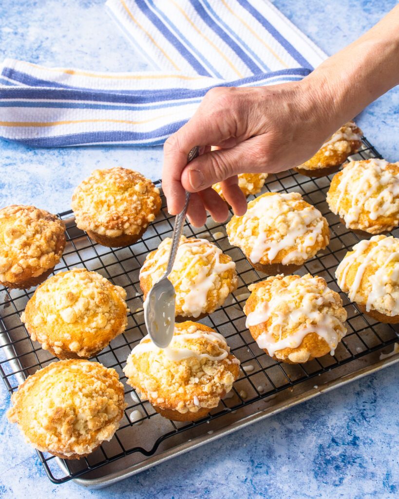 A hand drizzling lemon glaze on lemon streusel muffins in a muffin pan.