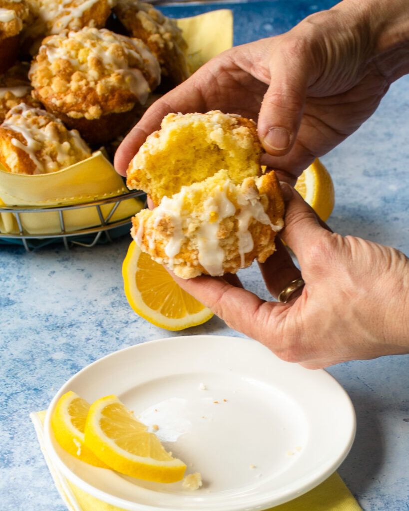 A hand breaking open a lemon streusel muffin over a white plate with lemon slices on it.