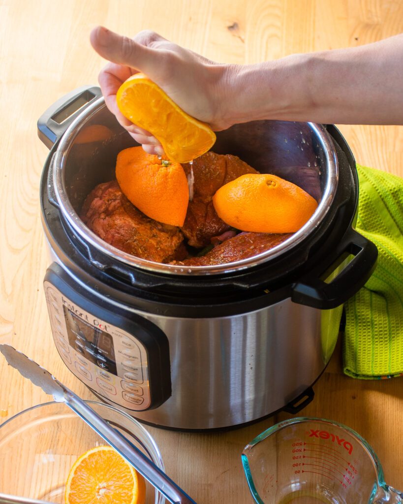 A hand squeezing half an orange into a pressure cooker with seasoned pork inside.