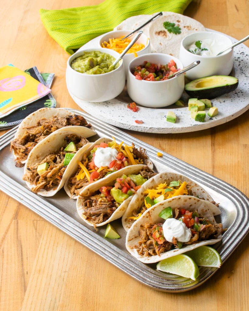 A silver tray with pork carnitas tacos on a wooden table with a tray of taco fixings in the background.