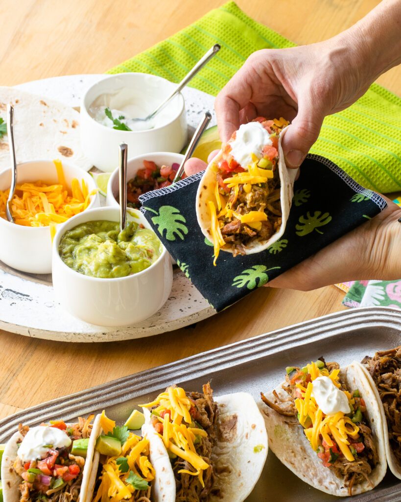 A hand holding a pork carnitas taco over a silver tray of more tacos.