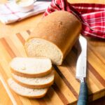 A loaf of white sandwich bread with a few slices cut off on a wooden cutting board with a bread knife.