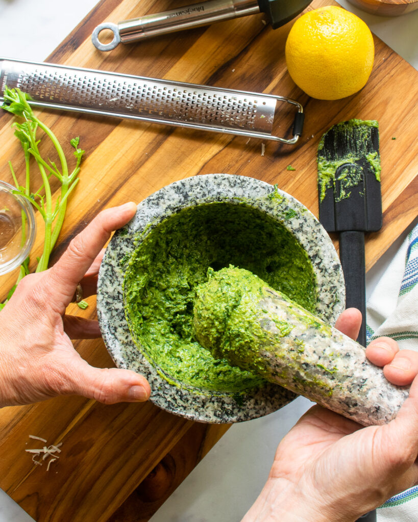Hands smashing basil pesto with a pestle in a mortar.