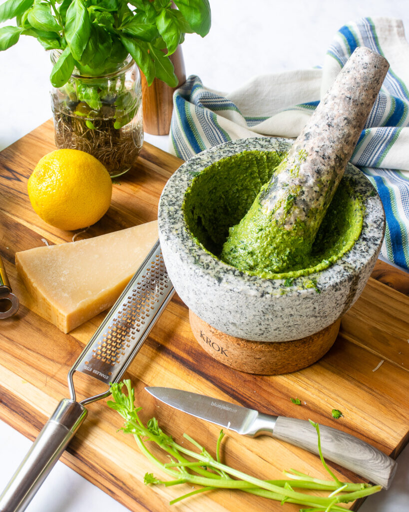 A stone mortar and pestle with basil pesto inside on a cutting board with some parm, a lemon and some basil.
