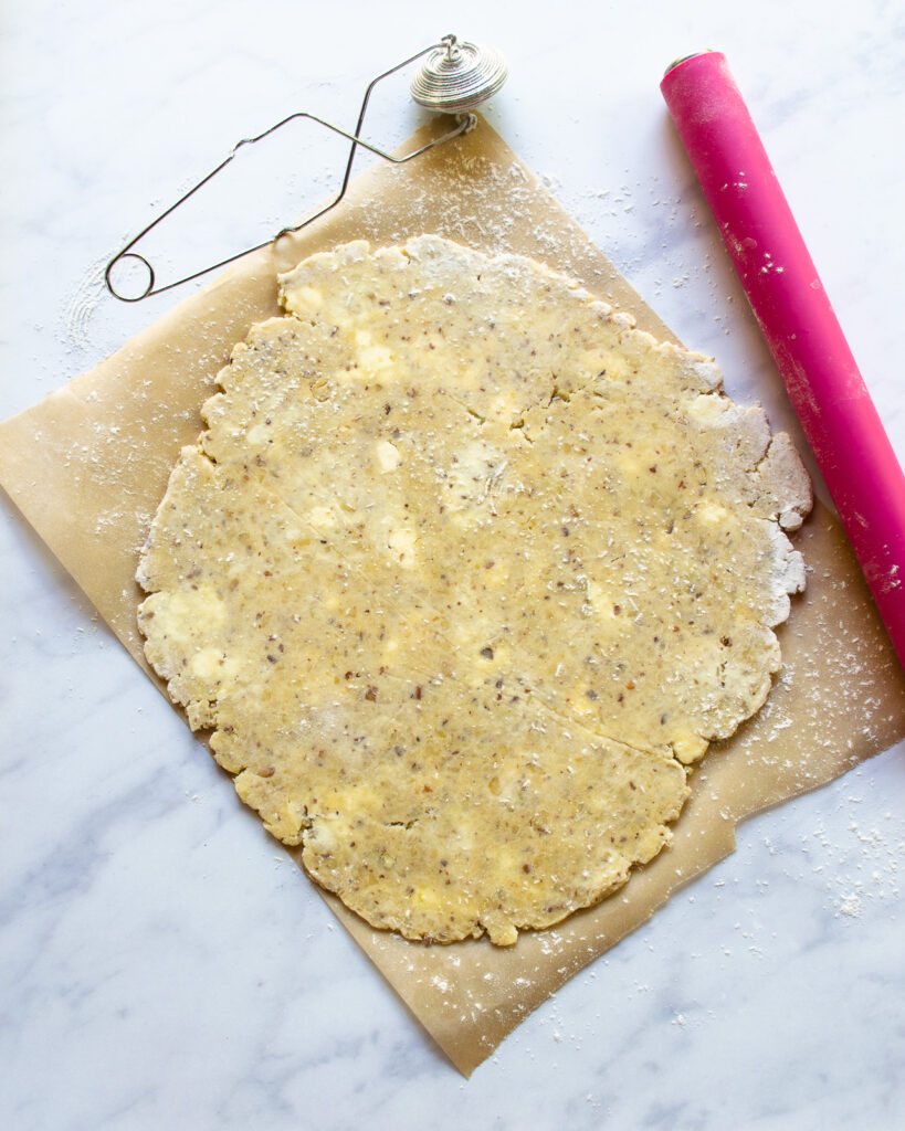 Pastry rolled out onto a piece of parchment paper with a pink rolling pin and a flour dusting wand on a marble counter.