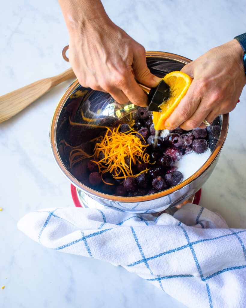 Hands squeezing orange juice into a bowl with frozen cherries, orange zest, and sugar, with a blue and white windowpane towel near by.