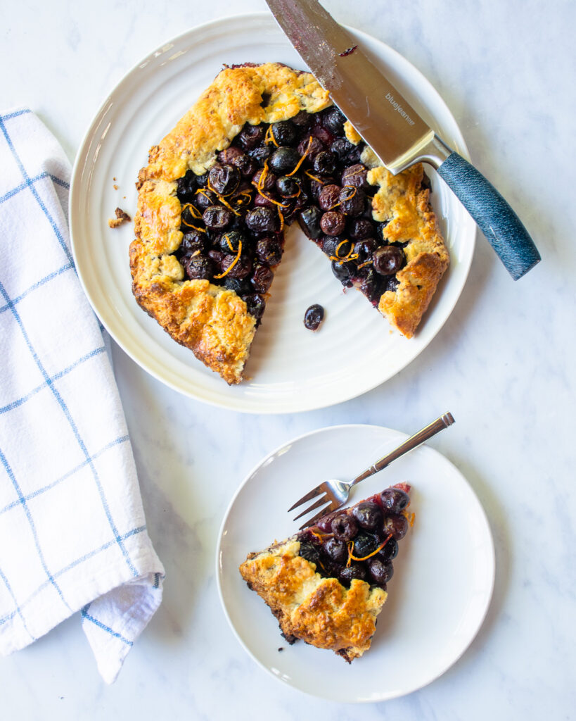 A cherry galette on a white plate with a knife resting on it and a piece cut out. The piece on a second white plate with a fork.