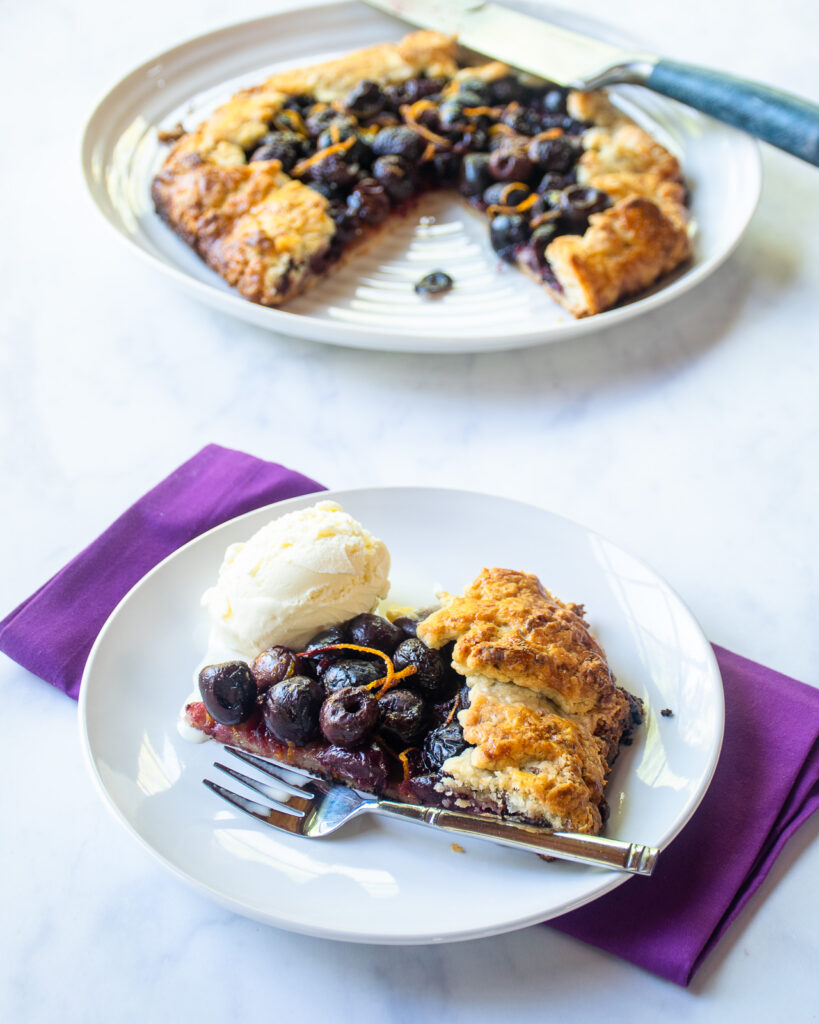 A piece of cherry galette and a scoop of vanilla ice cream on a white plate with a fork and a purple napkin. The rest of the tart in the background with a knife.