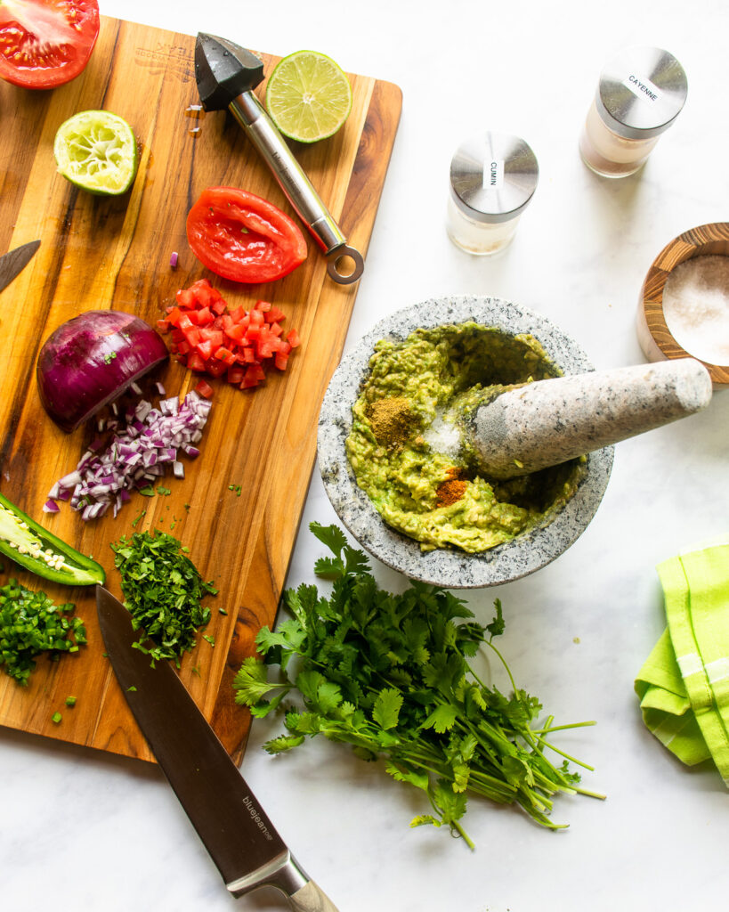 Looking down on a mortar with smashed avocado and some spices in it next to a cutting board with chopped tomato, red onion, cilantro and jalapeno pepper.