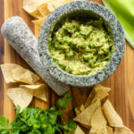 Looking down into a mortar of guacamole with tortilla chips and fesh cilantro near by.