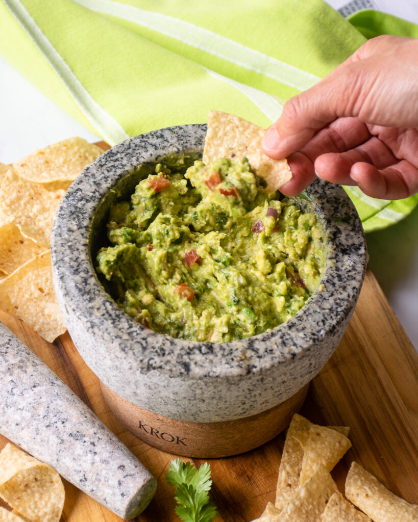 A hand scooping up guacamole on a tortilla chip from a mortar.