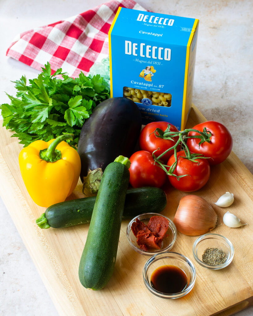 INgredients on a cutting board - a box of pasta, yellow pepper, zucchini, eggplant, vine tomatoes, onion, garlic, tomato paste, balsamic vinegar and dried herbs.