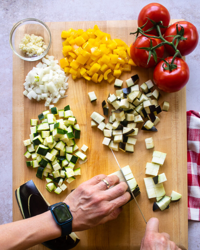 Hands chopping ingredients for ratatouille cavatappi on a cutting board - eggplant, zucchini, yellow pepper, tomatoes, etc...