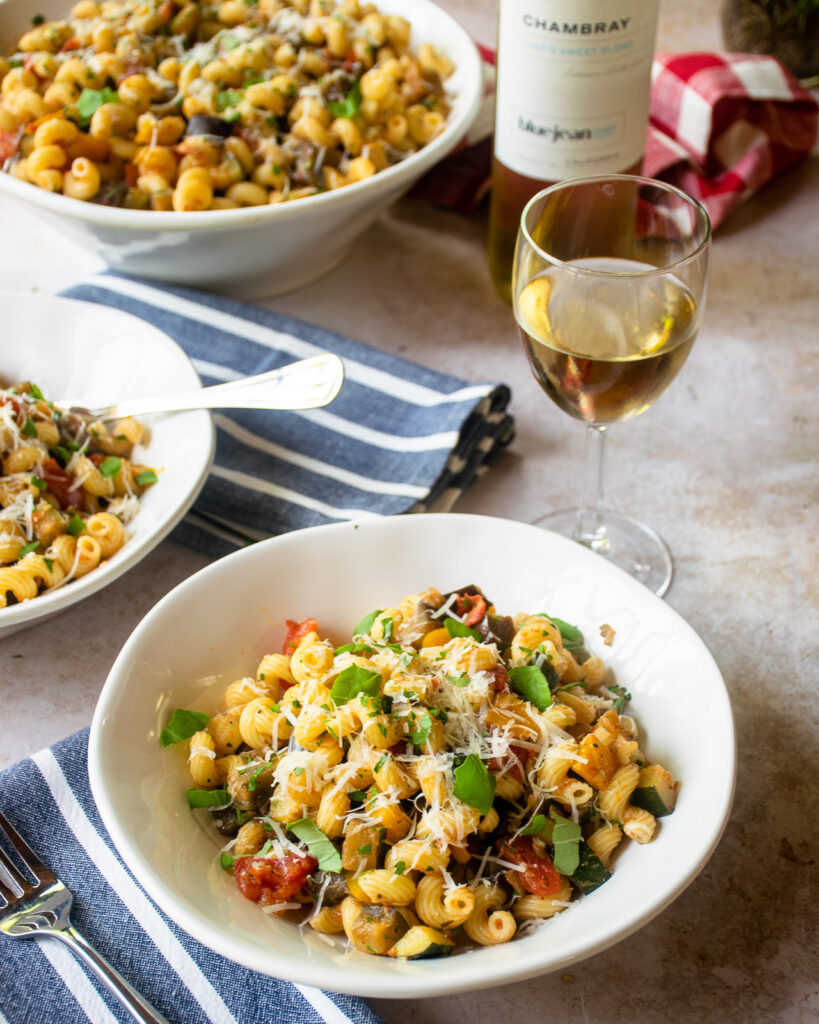 A white bowl with ratatouille cavatappi, blue and white striped napkins, a glass of wine with bottle in the background.