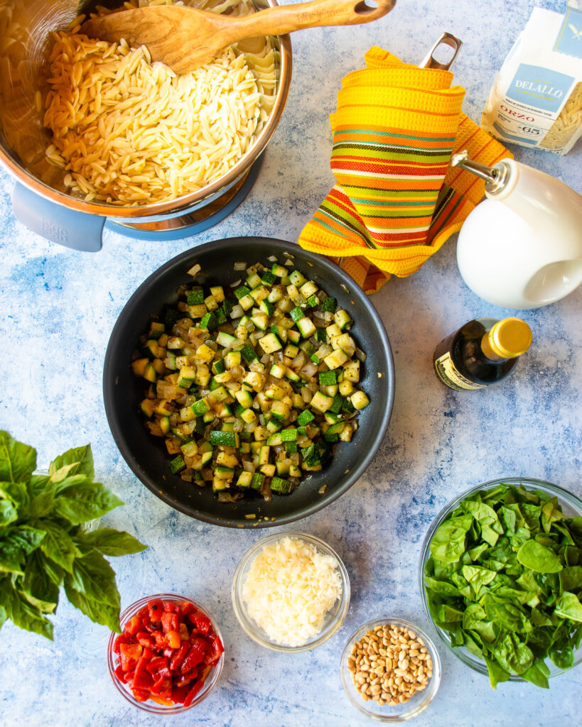 Looking down into a skillet with onion an zucchini next to a bowl of cooked orzo and some other ingredients - roasted red pepper, grated Parmesan cheese, toasted pinenuts and spinach.