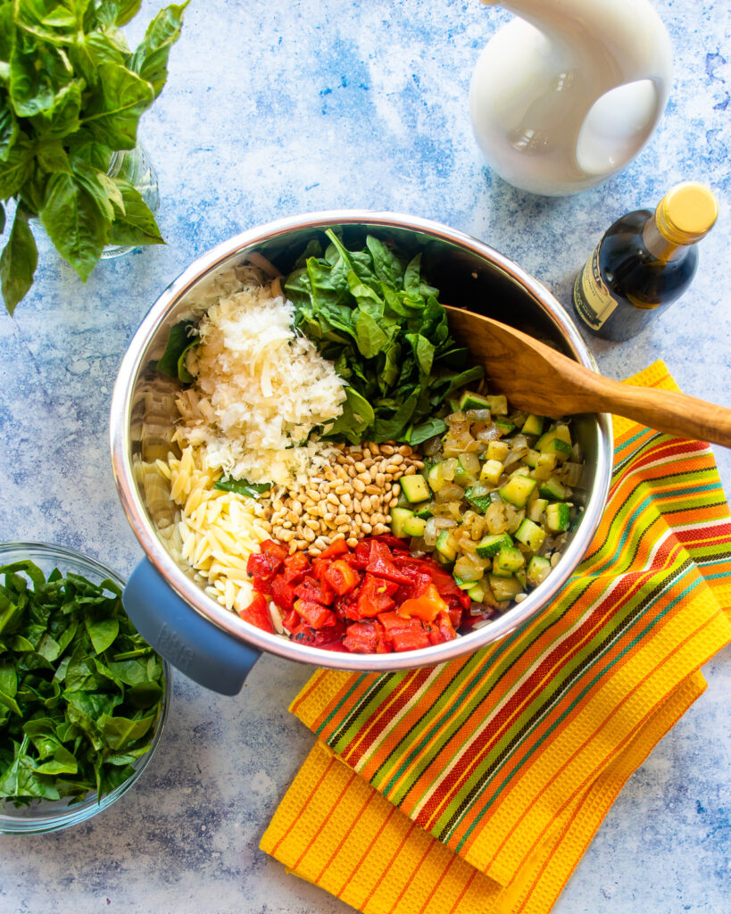 Looking down into a bowl with all the ingredients for a summer orzo salad, with a yellow striped towel next to it.