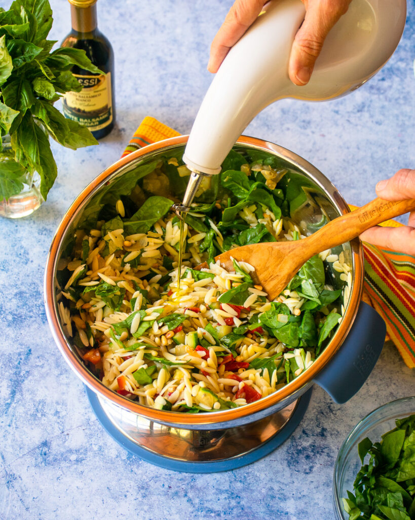 Pouring olive oil into a summer orzo salad with a wooden spoon sticking out of a stainless steel bowl.