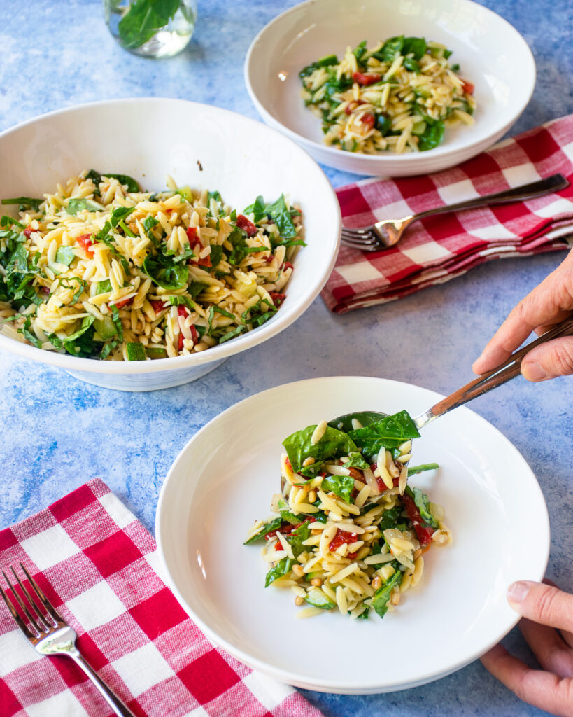 Spooning summer orzo salad out onto white plates with red and white checkered napkins and forks on the table.