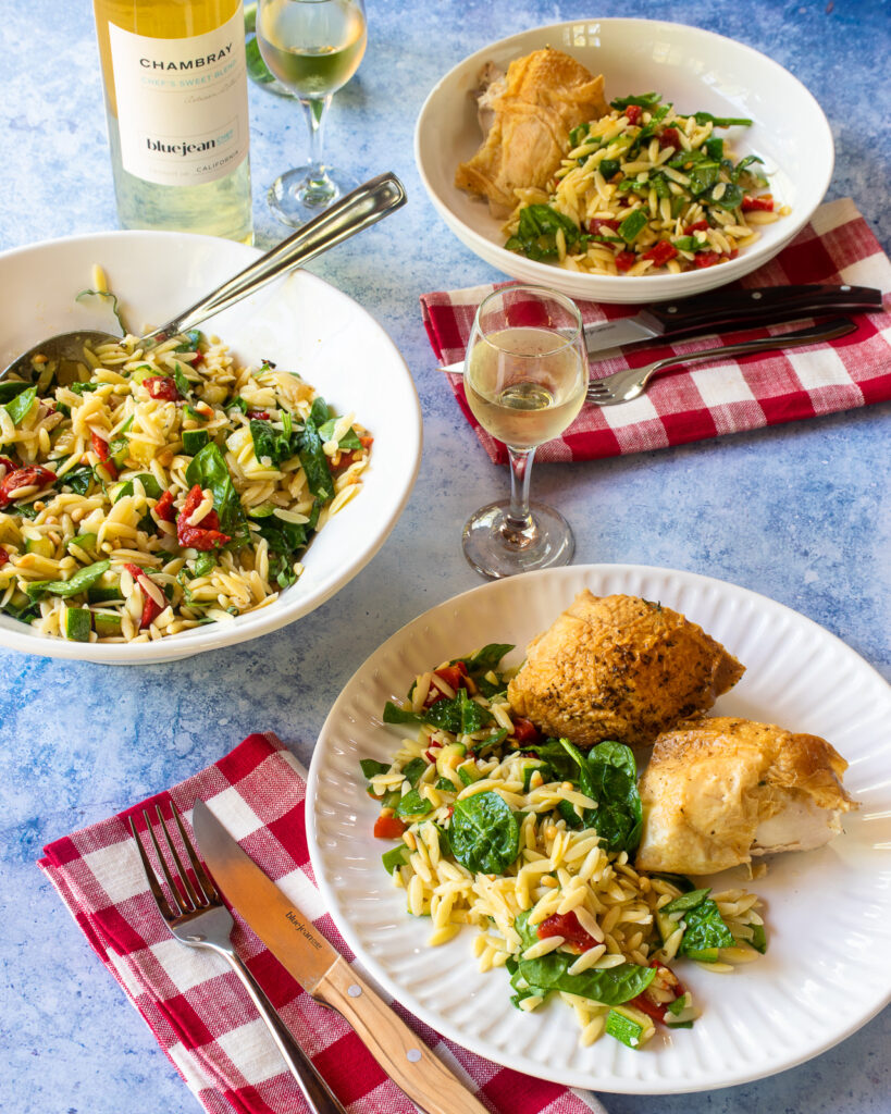 A table setting with a large bowl of summer orzo salad, two plates of chicken with the salad and some white wine.