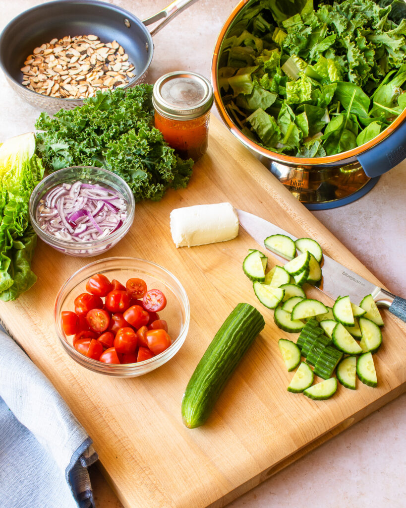 Salad ingredients on a cutting board with a bowl of greens near by.