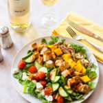 Grilled Chicken Salad with Mango on a white plate with a glass of white wine and a yellow napkin.