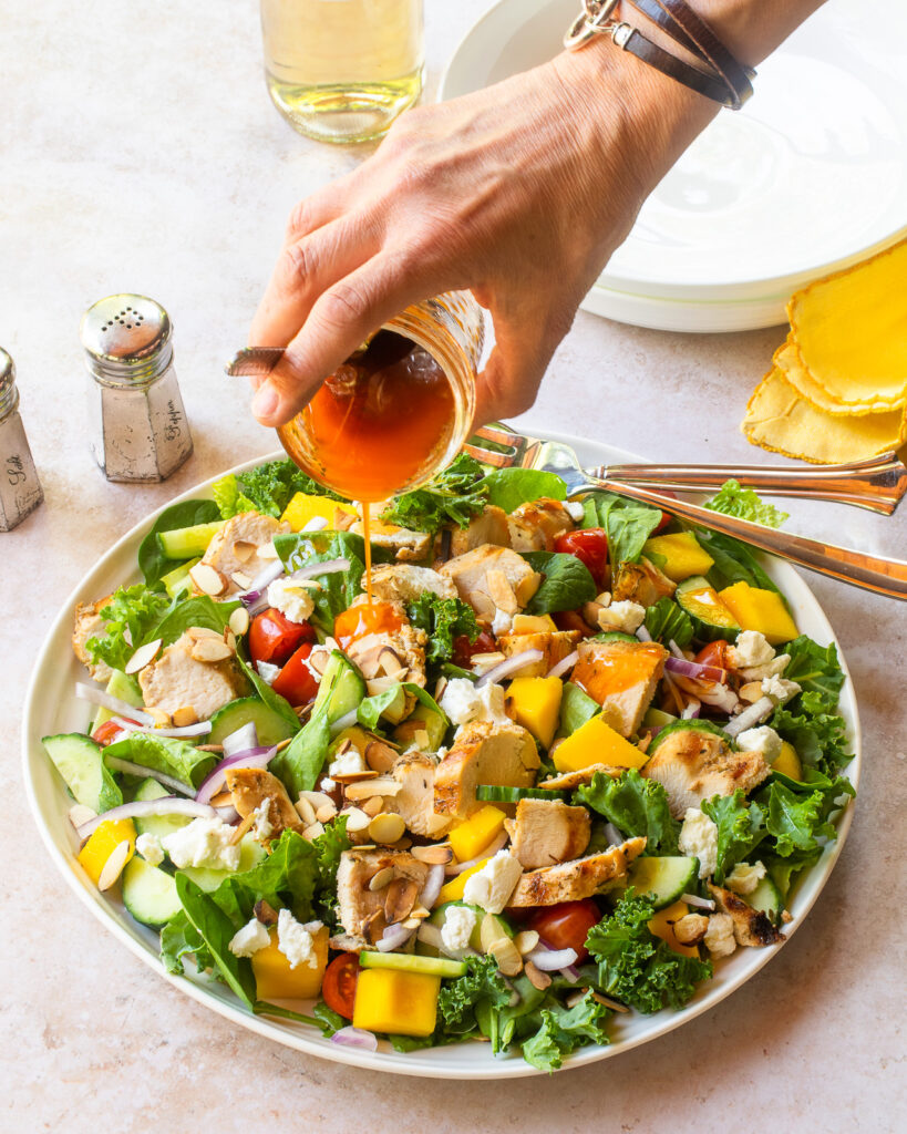 A hand pouring dressing over a grilled chicken salad on a white plate.