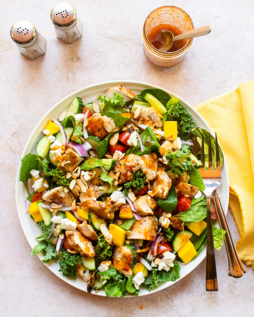 Looking down on a grilled chicken salad with mixed greens and mango on a white plate with yellow napkin.