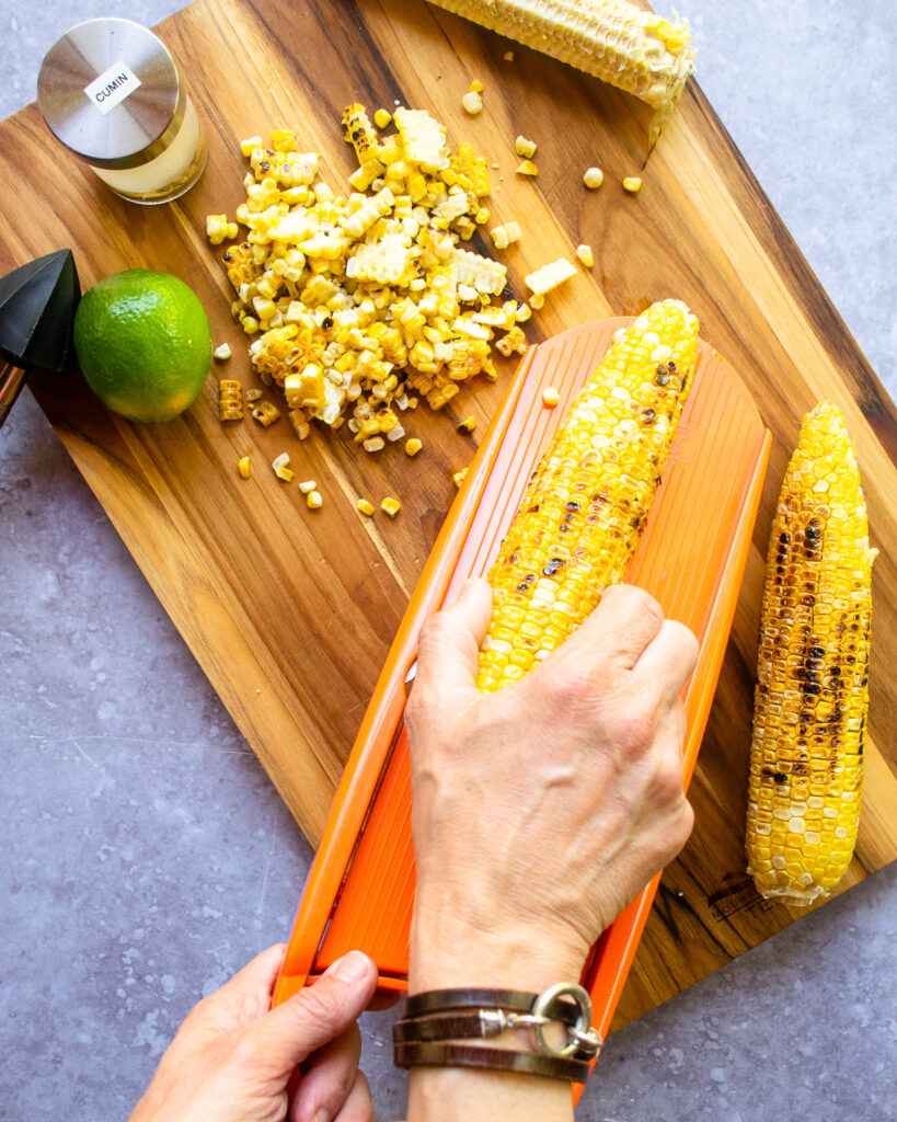 Hands slicing grilled corn kernels off the cob with a mandolin onto a cutting board.