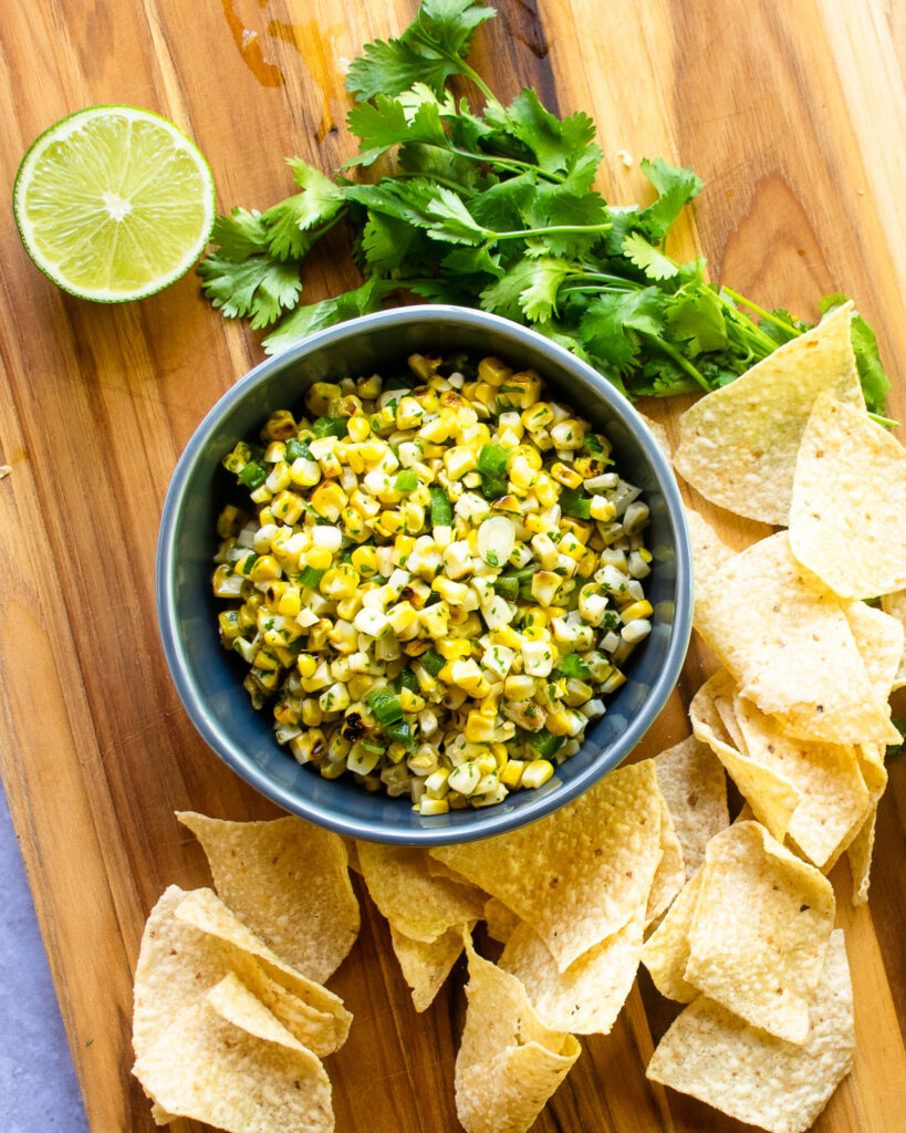 A bowl of corn and scallion salsa on a cutting board with chips and a lime half.