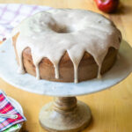 Apple Spice Cake on a cake stand with icing dripping down the sides and apples in the background.