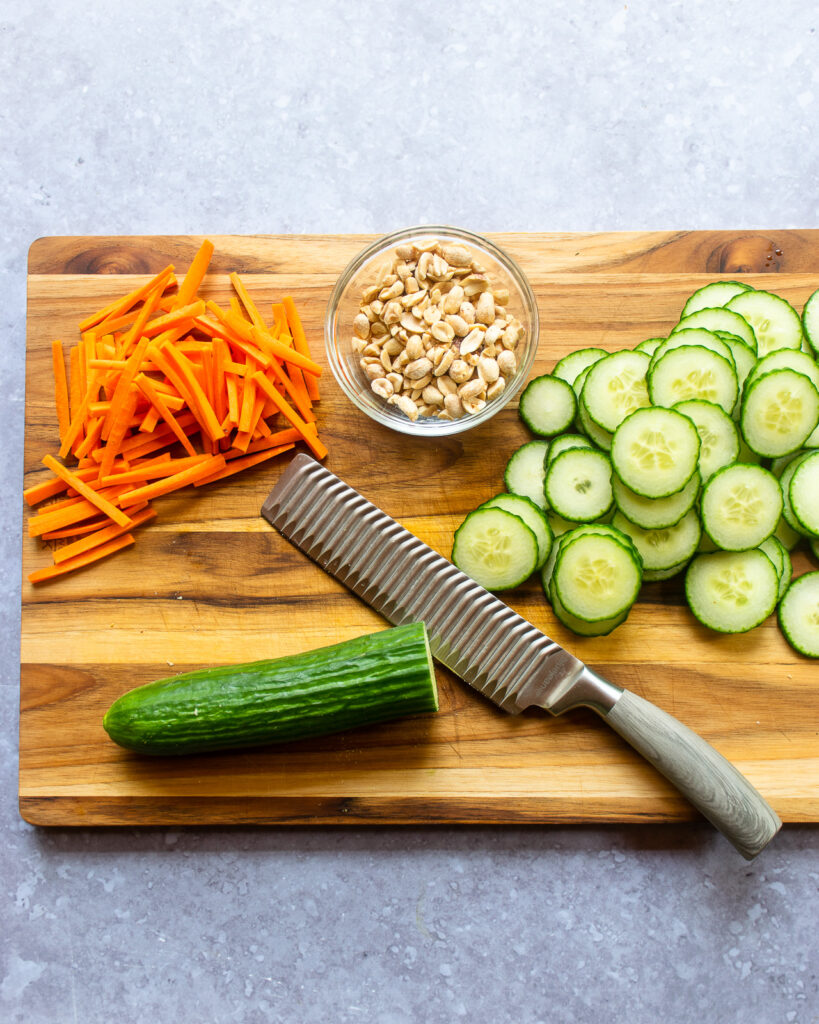 A nakiri knife with sliced cucumber, julienne carrots and peanuts on a cutting board.