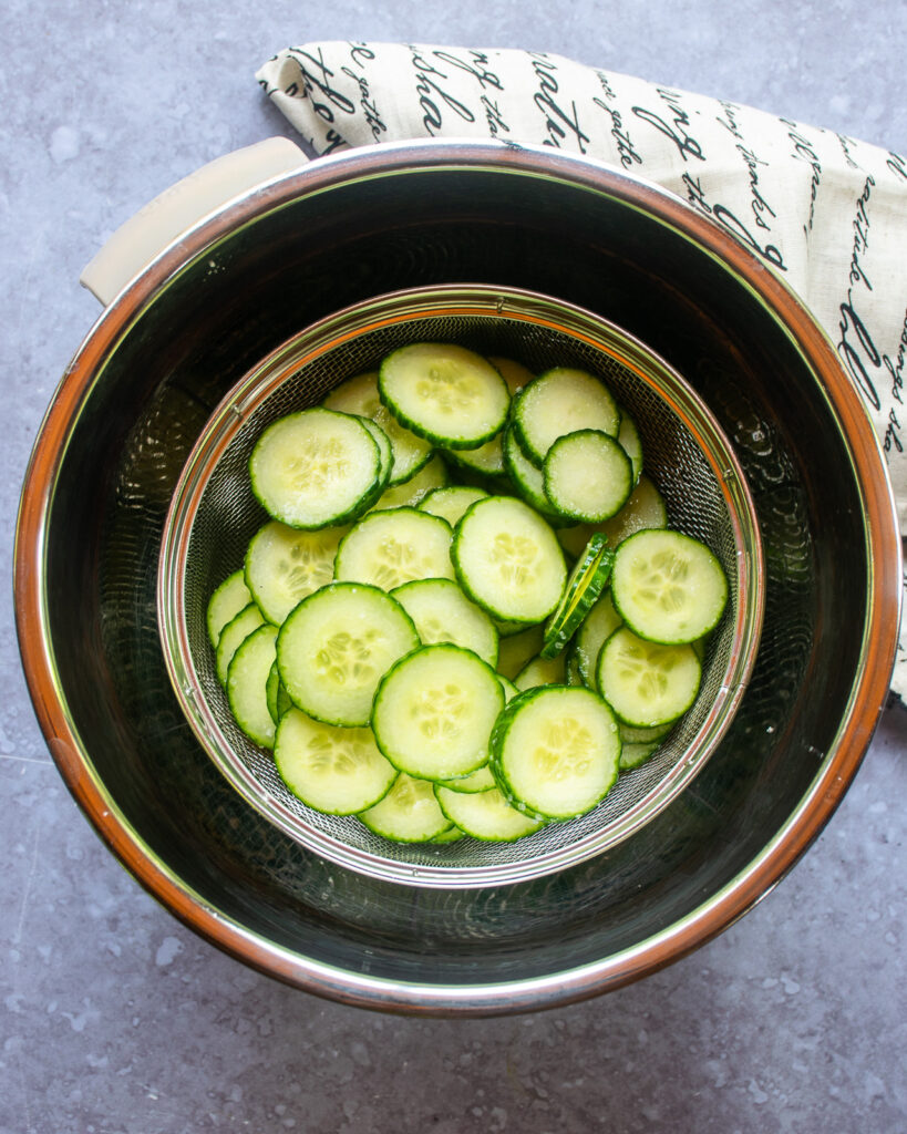 Cucumber slices salting in a colander in a stainless steel bowl.