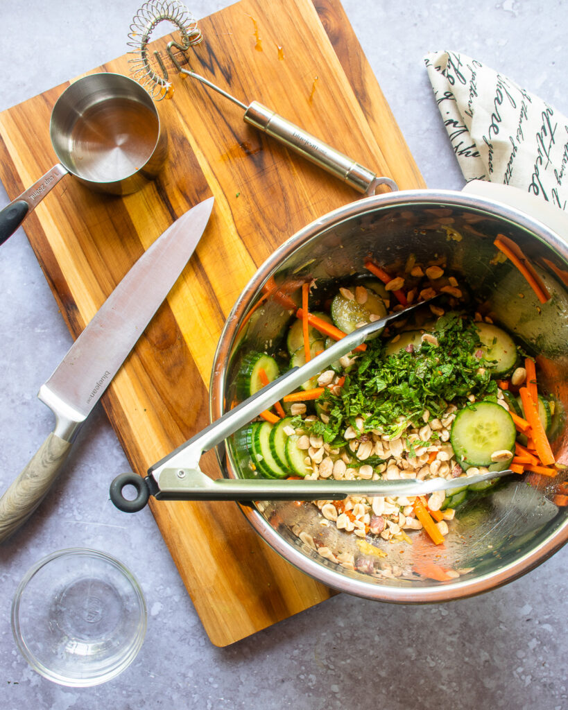 Looking down into a stainless steel bowl with Asian Cucumber Salad ingredients and a pair of tongs next to a chef's knife, measuring cup and whisk.