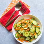 Looking down on a white bowl with Asian Cucumber Salad next to a red and an orange and white napkin, spoon and fork.