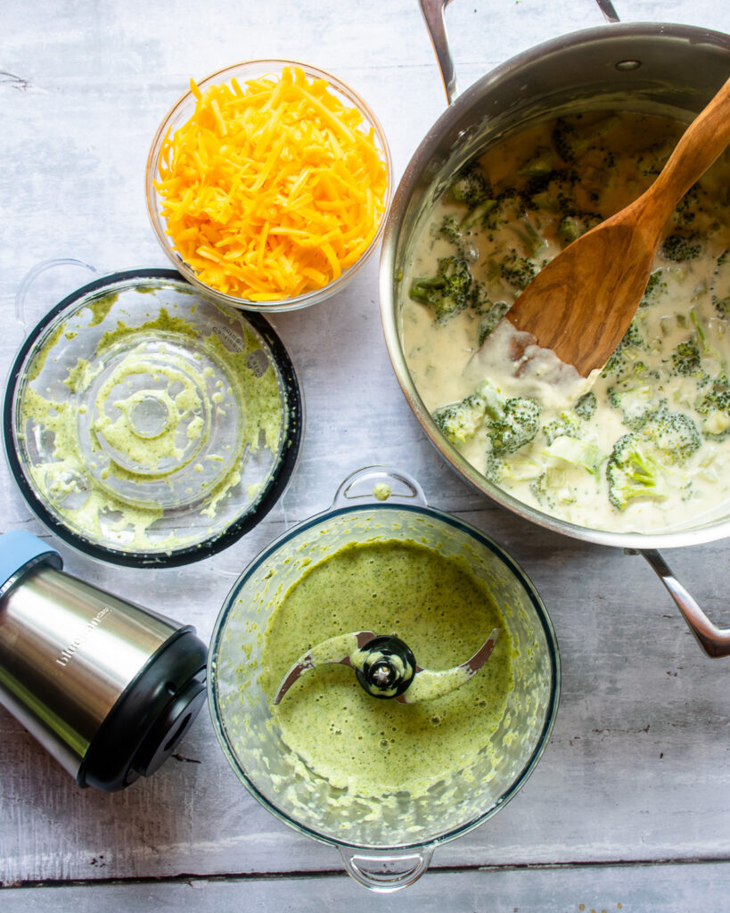 Looking down at a chopper with a green broccoli soup purée, a bowl of Cheddar cheese and a stockpot with cooked broccoli soup inside.