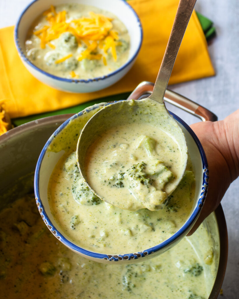 A close up of a ladle of chunky broccoli Cheddar soup being transfered from pot to a bowl and a second bowl of broccoli Cheddar soup in the background.