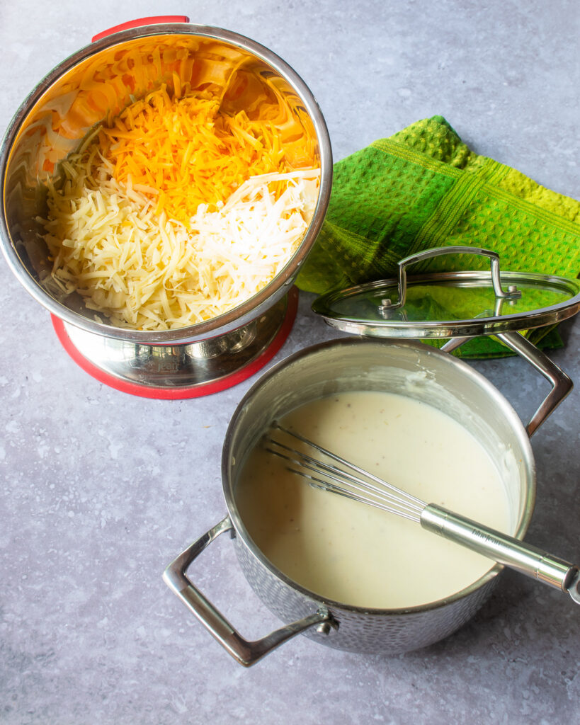 A saucepan of cream sauce with a whisk next to a bowl of grated cheeses and a green kitchen towel.