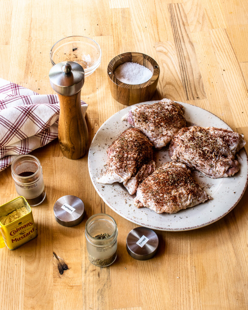 Chicken thighs seasoned with a spice rub on a plate with various seasonings around it.