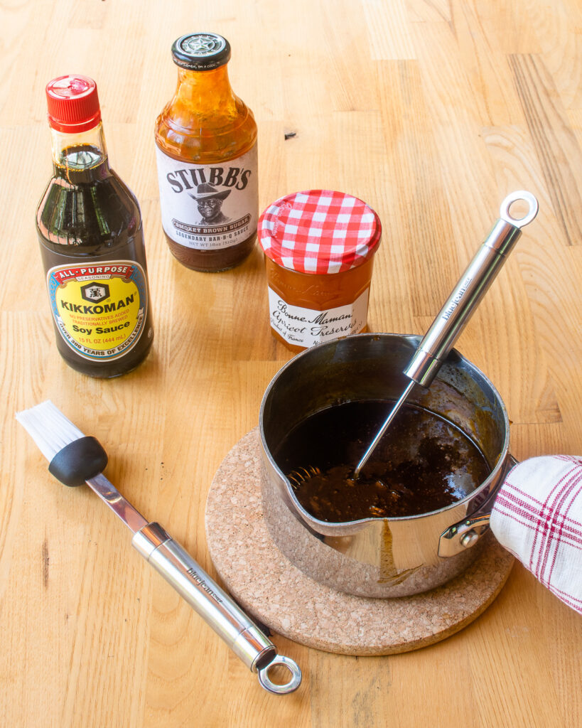 Ingredients for a BBQ sauce on a butcher block next to a small saucepan with whisk and basting brush.