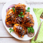 Looking straight down at a white oval platter with BBQ grilled chicken thighs and red onion, next to a green and white napkin with large fork.