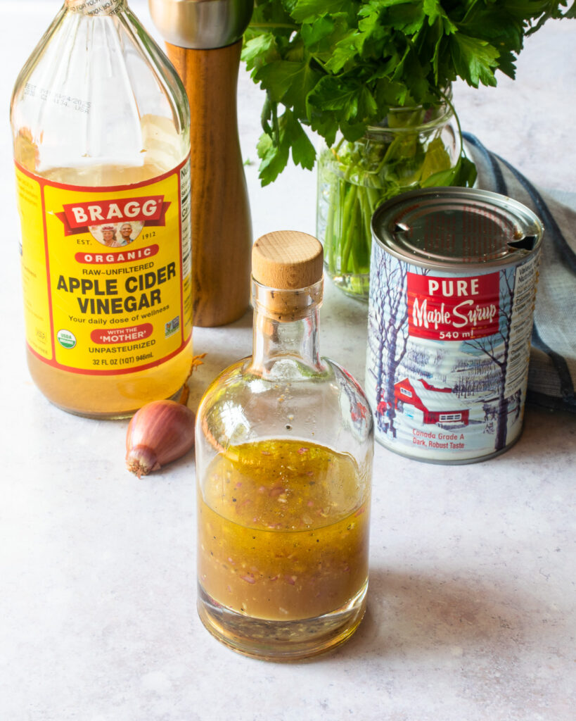 A bottle of vinaigrette with a bottle of apple cider vinegar, shallot and a can of maple syrup behind it.