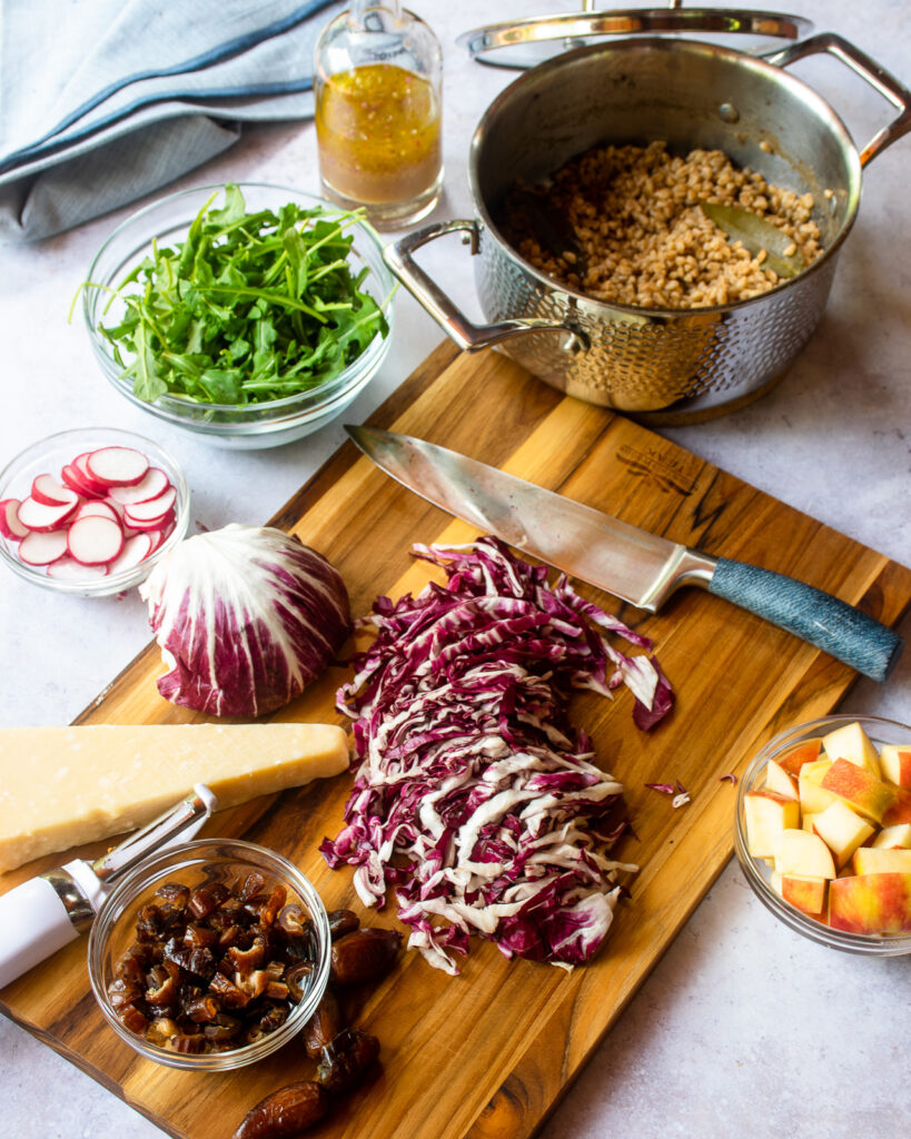 Ingredients for farro salad on a cutting board - radicchio, a saucepan of farro, arugula, apples, radishes, parmesan cheese and dates.