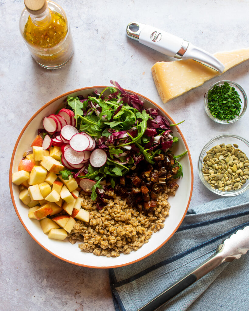 A farro salad with apples and arugula in a large serving bowl, arranged in sections, with a few other ingredients around the side.