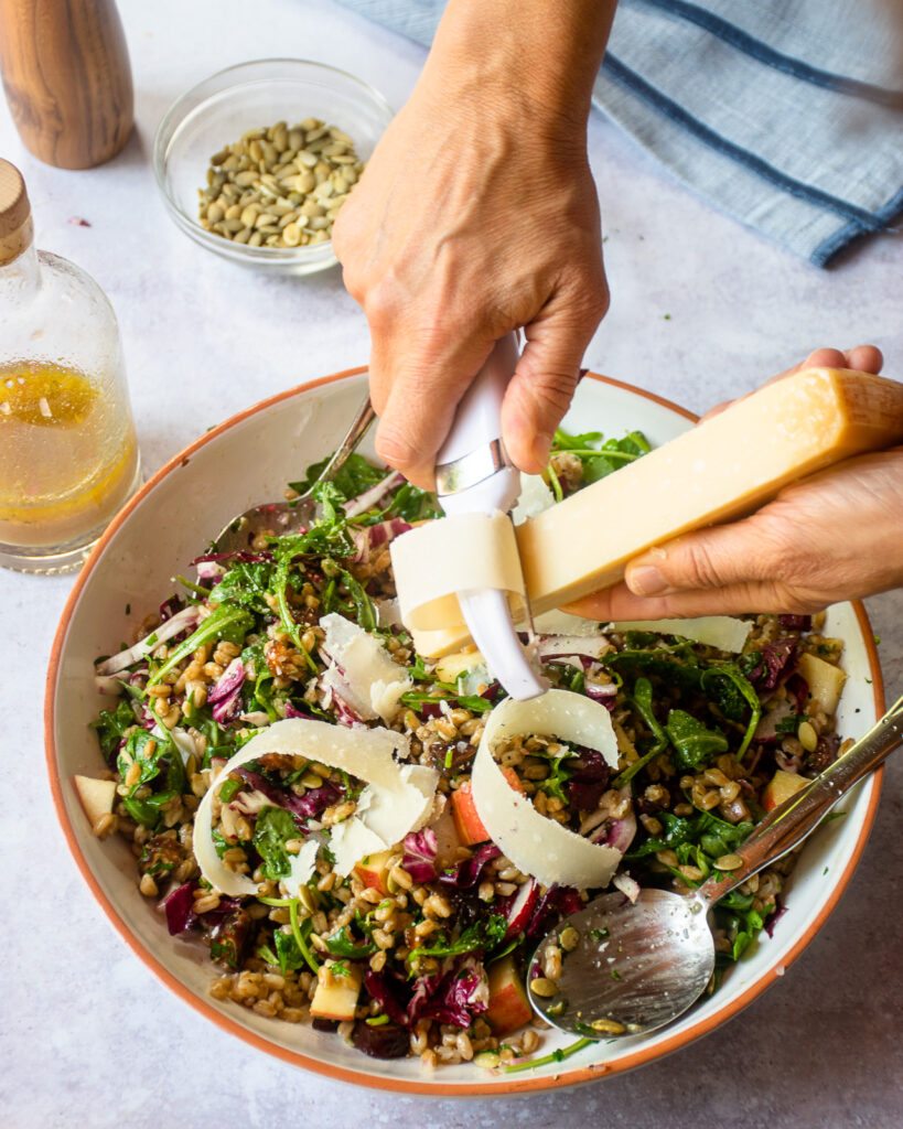 Hands adding peelings of Parmesan cheese to the top of a farro salad.